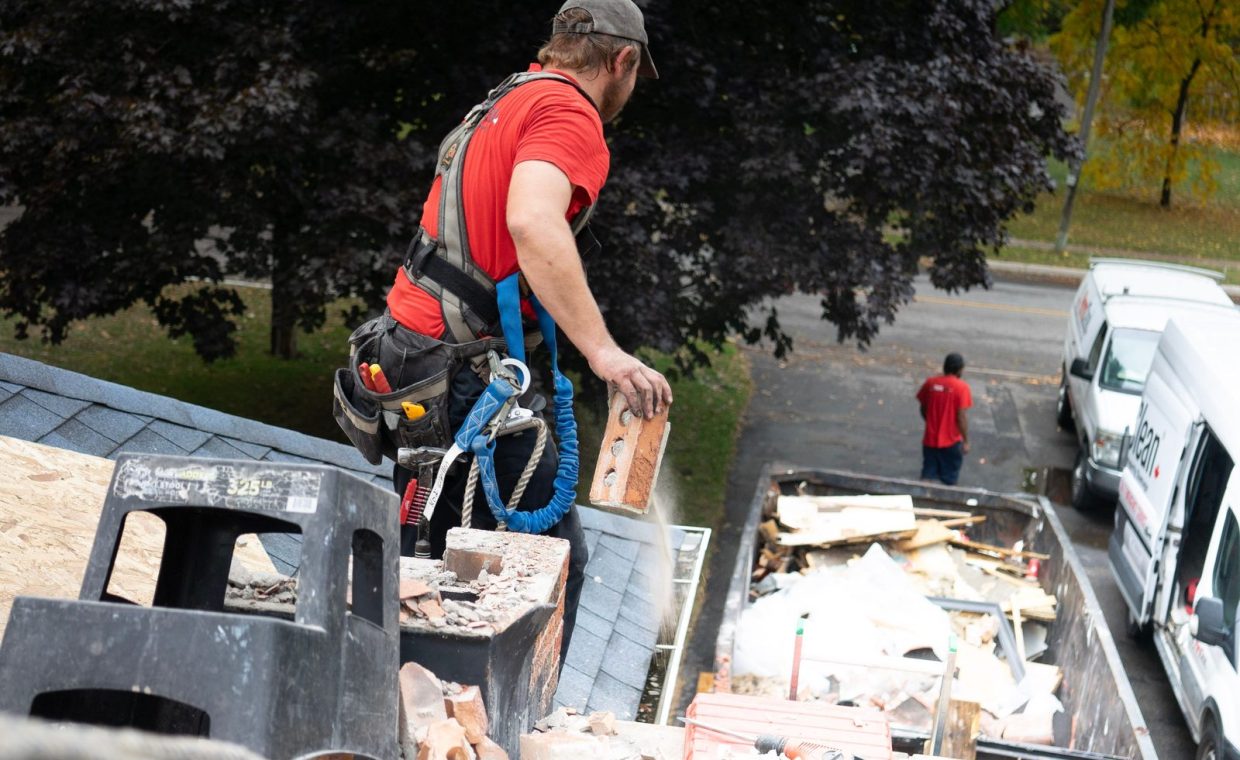Worker in safety harness removes bricks from roof into dumpster below.