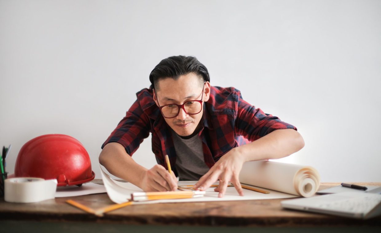 Inspired young male engineer in glasses drawing draft on paper bending over table against white wall