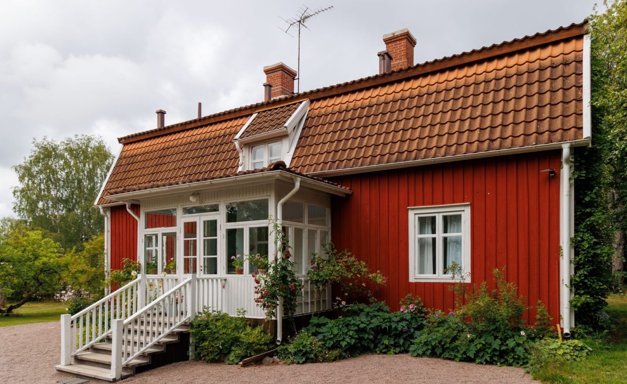 A picturesque rural red wooden house with white trim and a tailed gable roof.