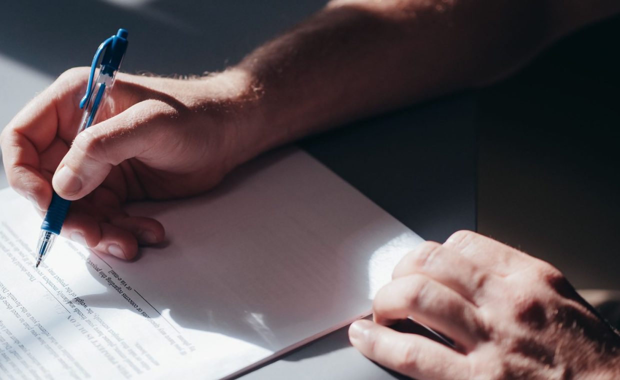 A close-up image of a person's hand signing a document with a blue pen in natural lighting.
