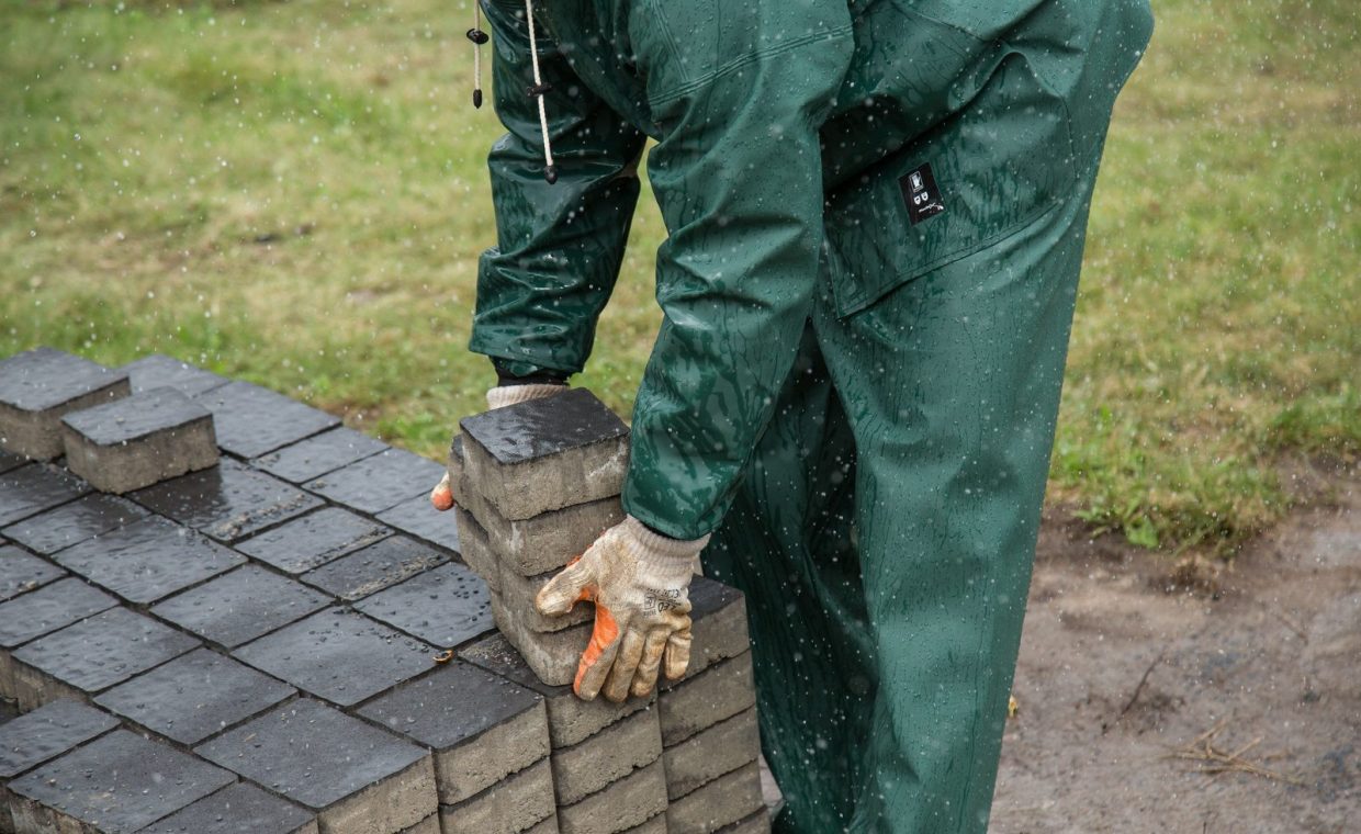 A worker in rain gear carefully places paving stones during a rainy day outdoor construction project.