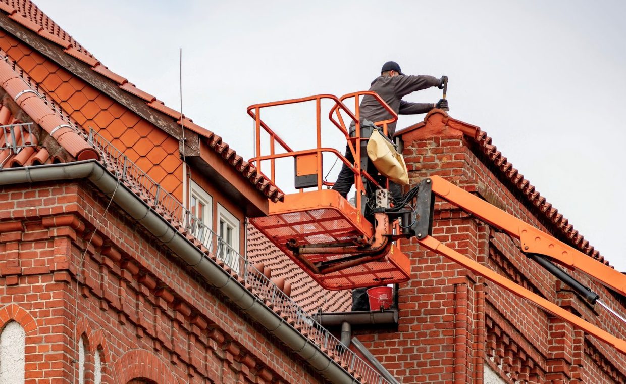 Roofer in a cherry picker conducting maintenance on a red brick building with a tiled roof.