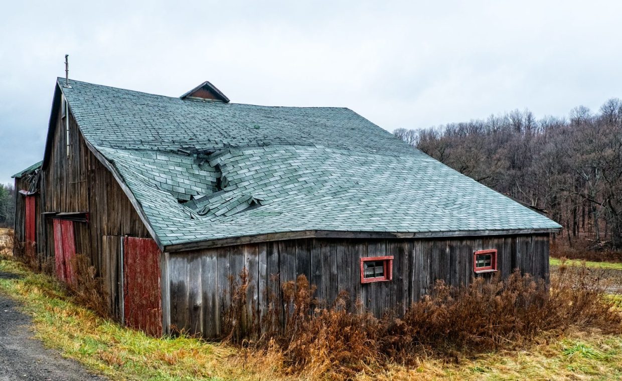 A weathered wooden barn with a damaged roof in a rural New York landscape.