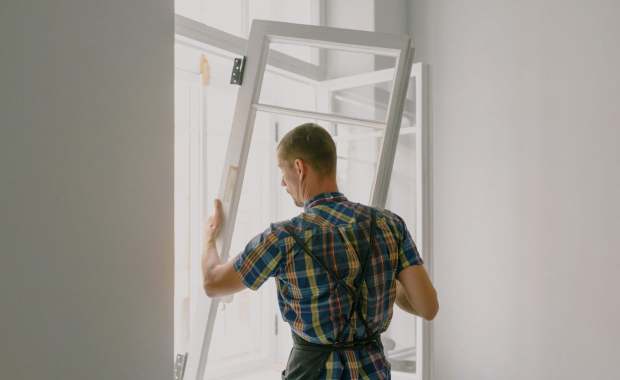 Back view of anonymous male fitter in checkered shirt and apron mounting window with hinges near white walls at home